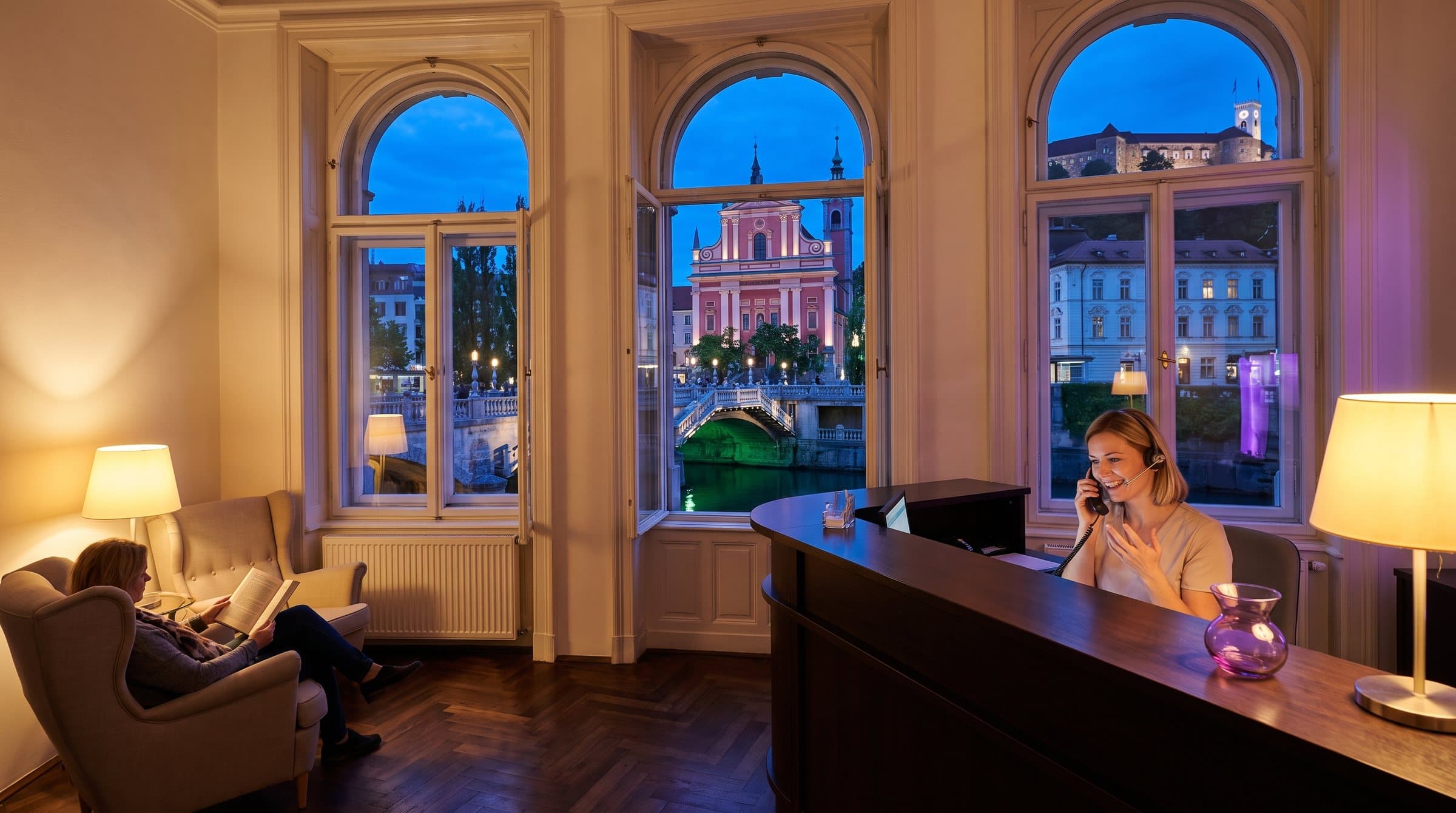 Receptorka s slušalkami se oglaša na recepciji zobozdravstvene ordinacije v secesijski stavbi v Ljubljani, za oknom Tromostovje, Ljubljanica in grad ob modri uri